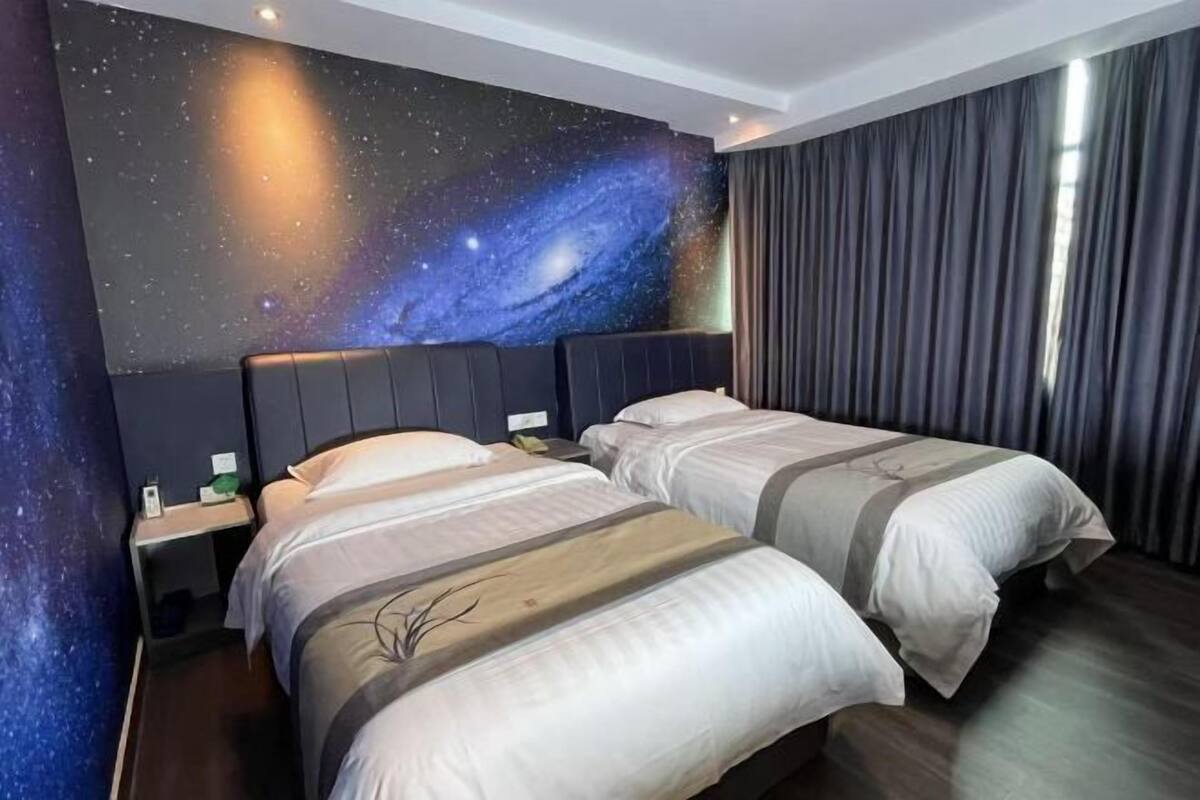 Habitación de hotel con dos camas, un mural en la pared con temática de galaxia y vistas del cielo nocturno a través de la ventana.