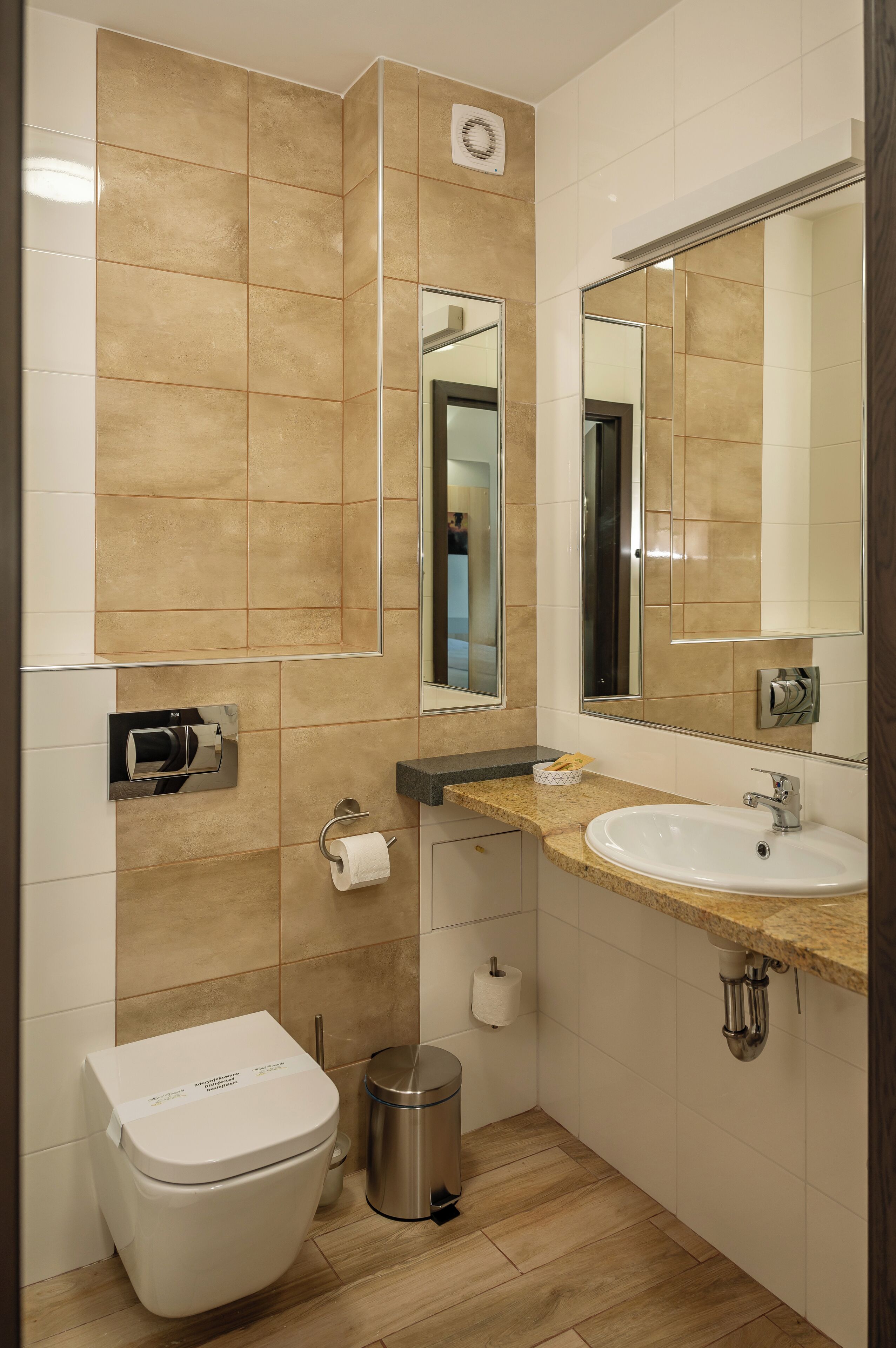 A modern bathroom with beige tiles, a white toilet, a sink with a mirror, and a trash can.