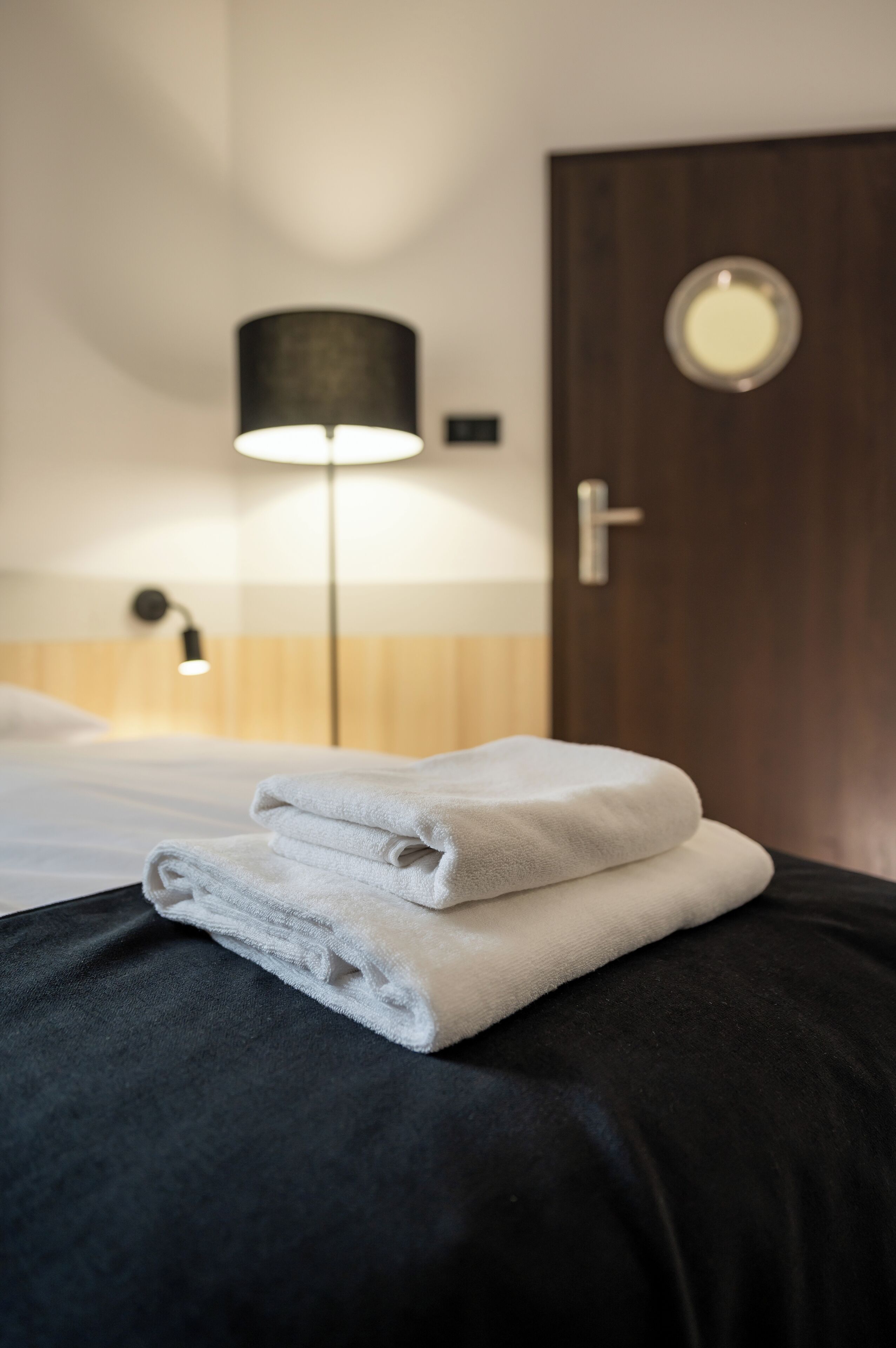 Un lit bien fait, avec des serviettes blanches pliées, une lampe noire et une porte en bois, dans une chambre d’hôtel.