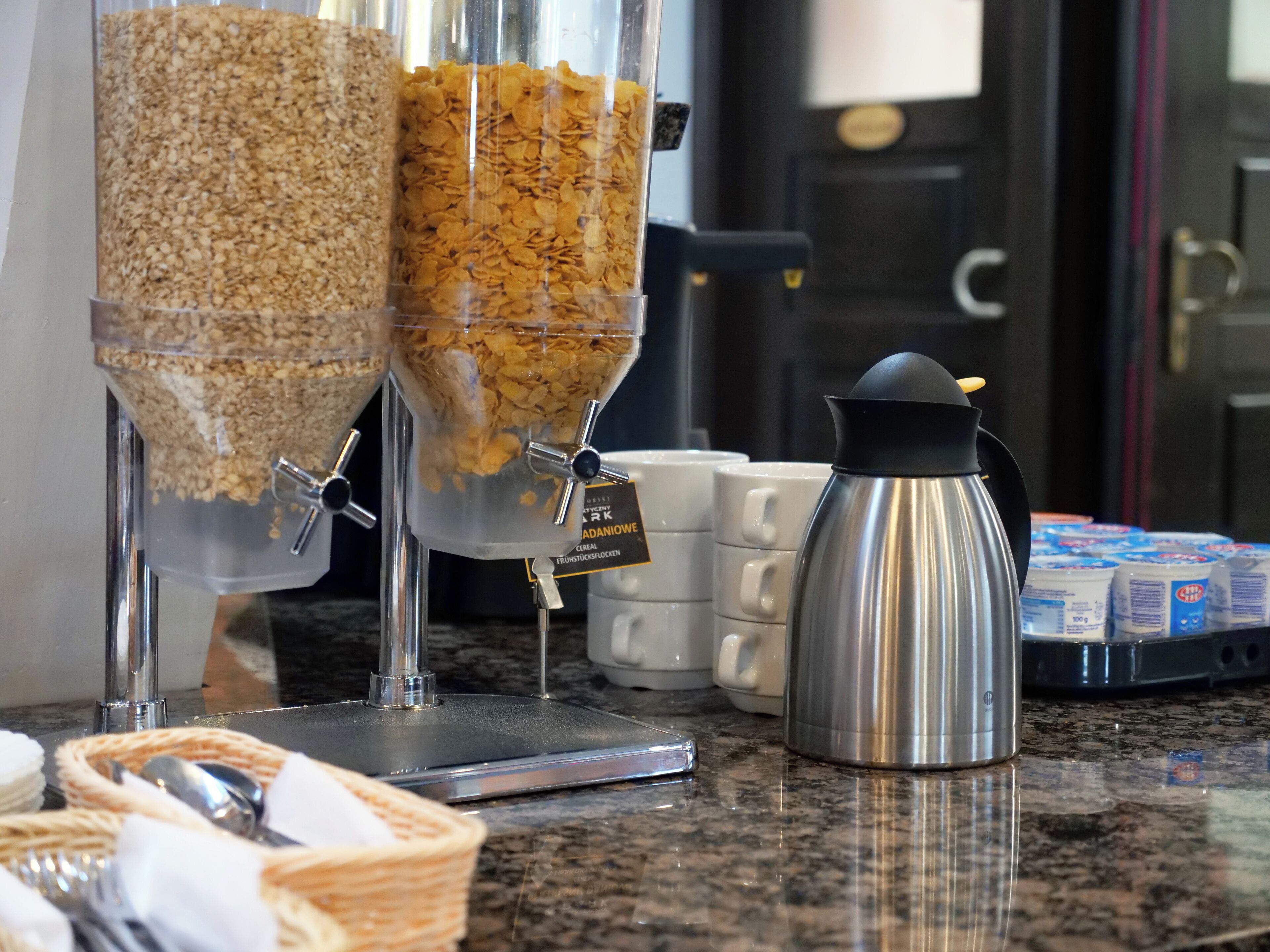 A breakfast buffet with cereal dispensers, a coffee pot, and milk cartons on a granite countertop.