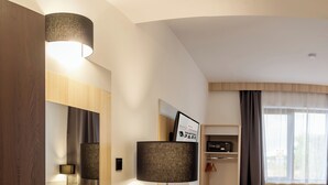 A hotel room with a bed, bedside table, lamp, desk, chair, and a view of the outside through the window.