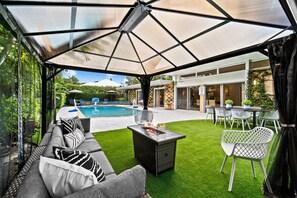 Un'area esterna coperta con piscina, un tavolo da pranzo con sedie e un divano con cuscini.