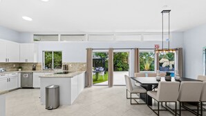 A modern kitchen with white cabinets, a central island, and a dining area with a table and chairs. - 4BR Intracoastal Retreat With Pool, Games & More! (West Palm Beach)