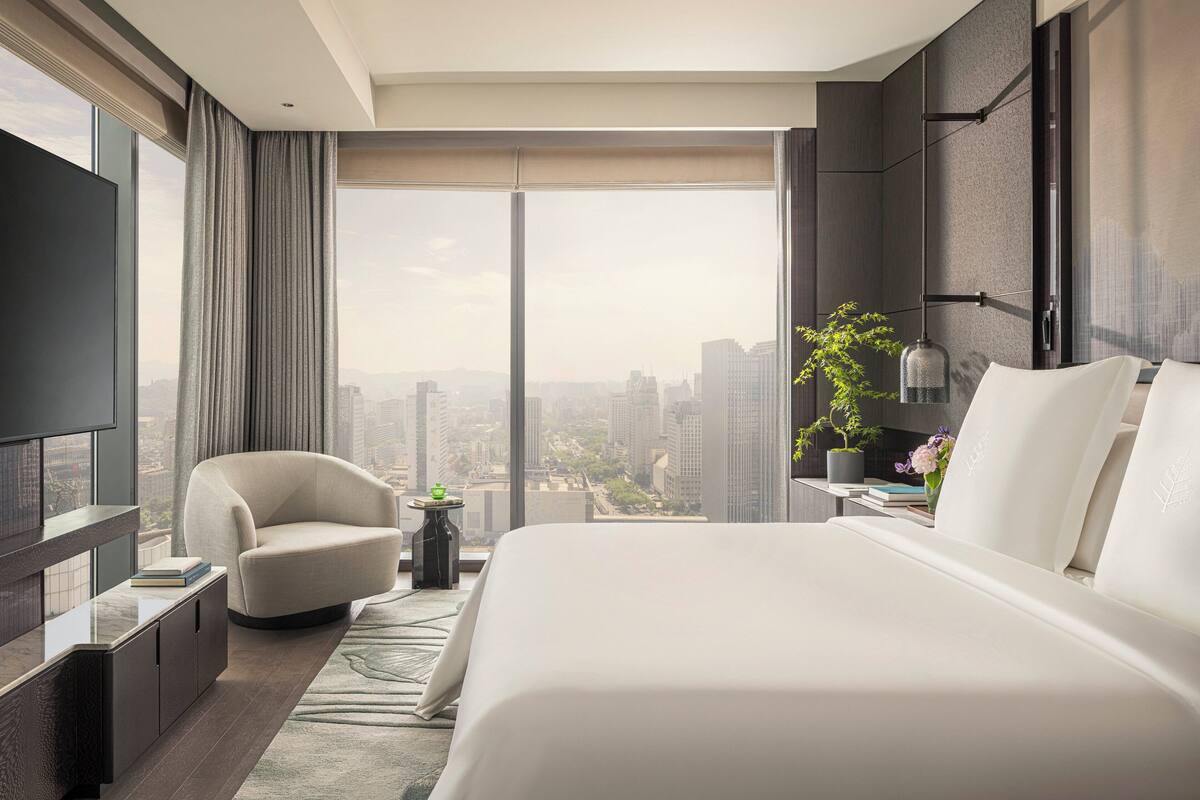 A modern hotel room with a large bed, a seating area, a TV, and a city view.