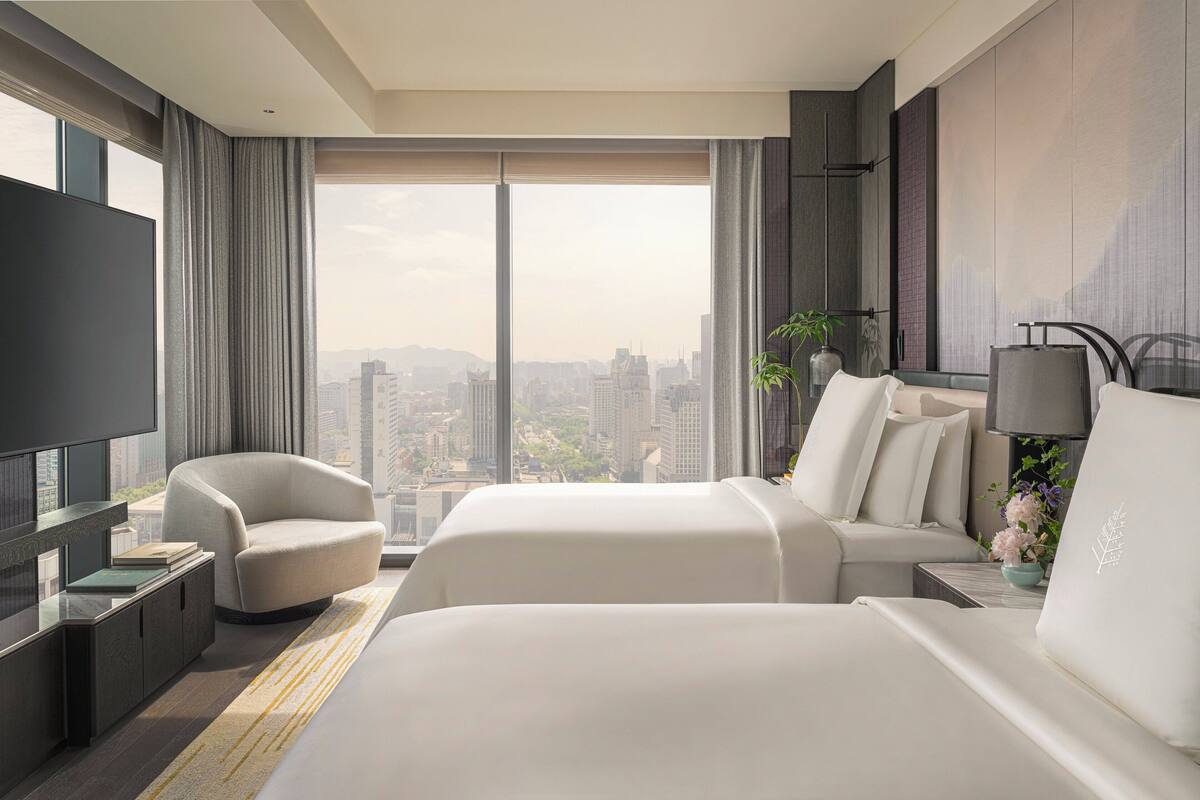 A modern hotel room with a large bed, a sofa, a TV, and a city view.