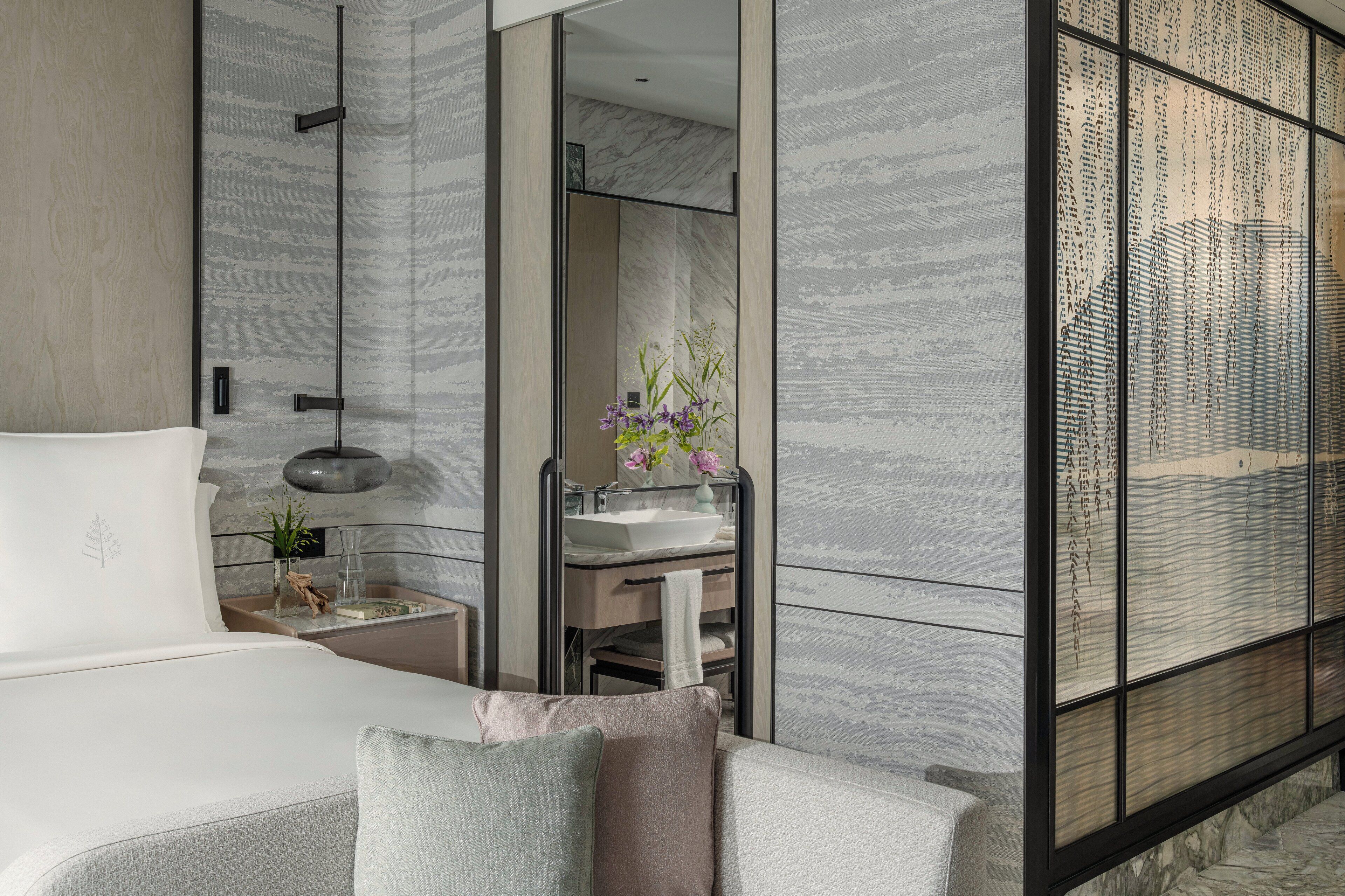 Un dormitorio moderno con una cama grande, una mesita de noche con lámpara, un baño con lavabo y espejo, y un panel decorativo en la pared.