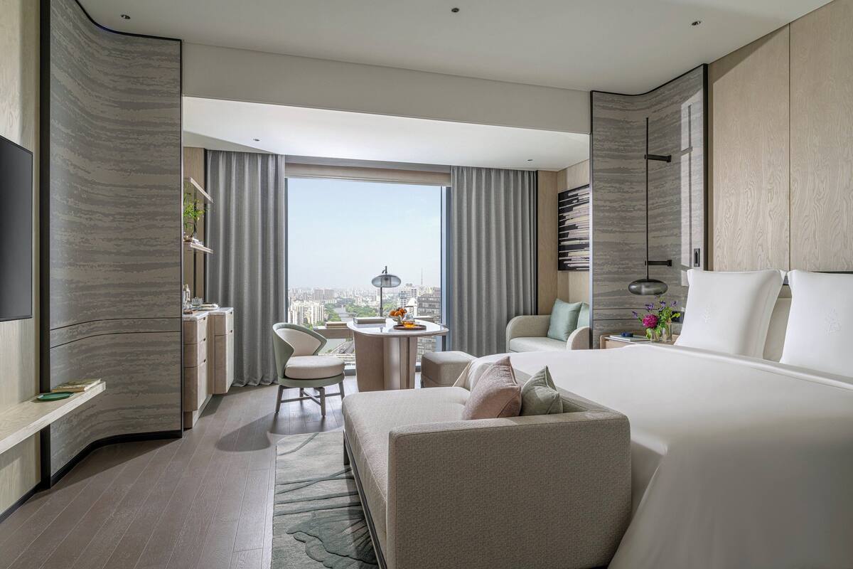 A modern hotel room with a large bed, a sitting area with a sofa and chairs, a small table with a lamp, and a view of the city through large windows.