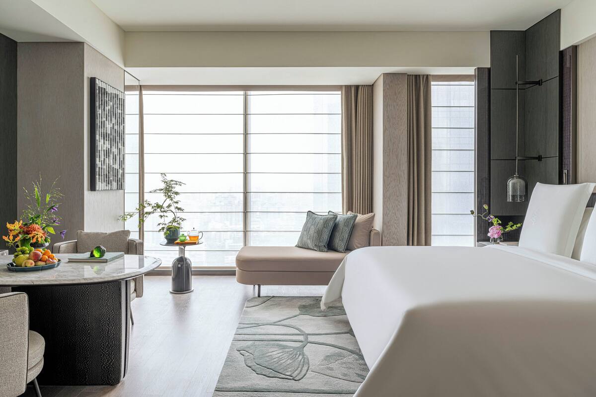 A modern hotel room with a large bed, a seating area, a dining table, and a view of the city through large windows.