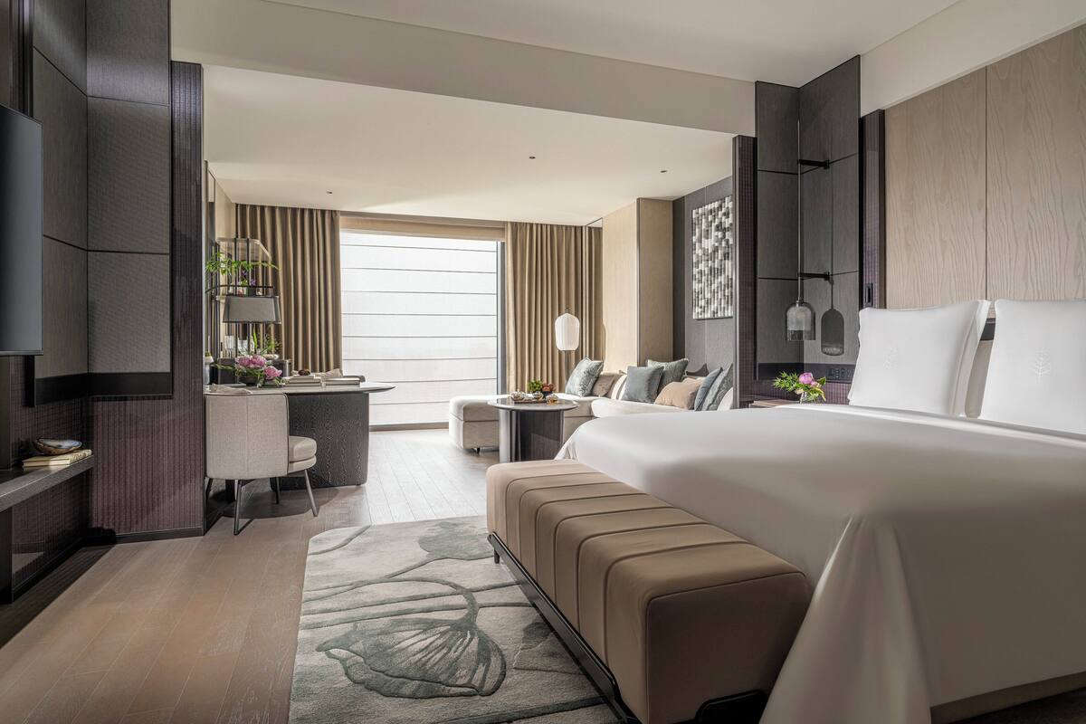 A modern hotel room with a large bed, a desk with a chair, a TV, and a view of the outside.