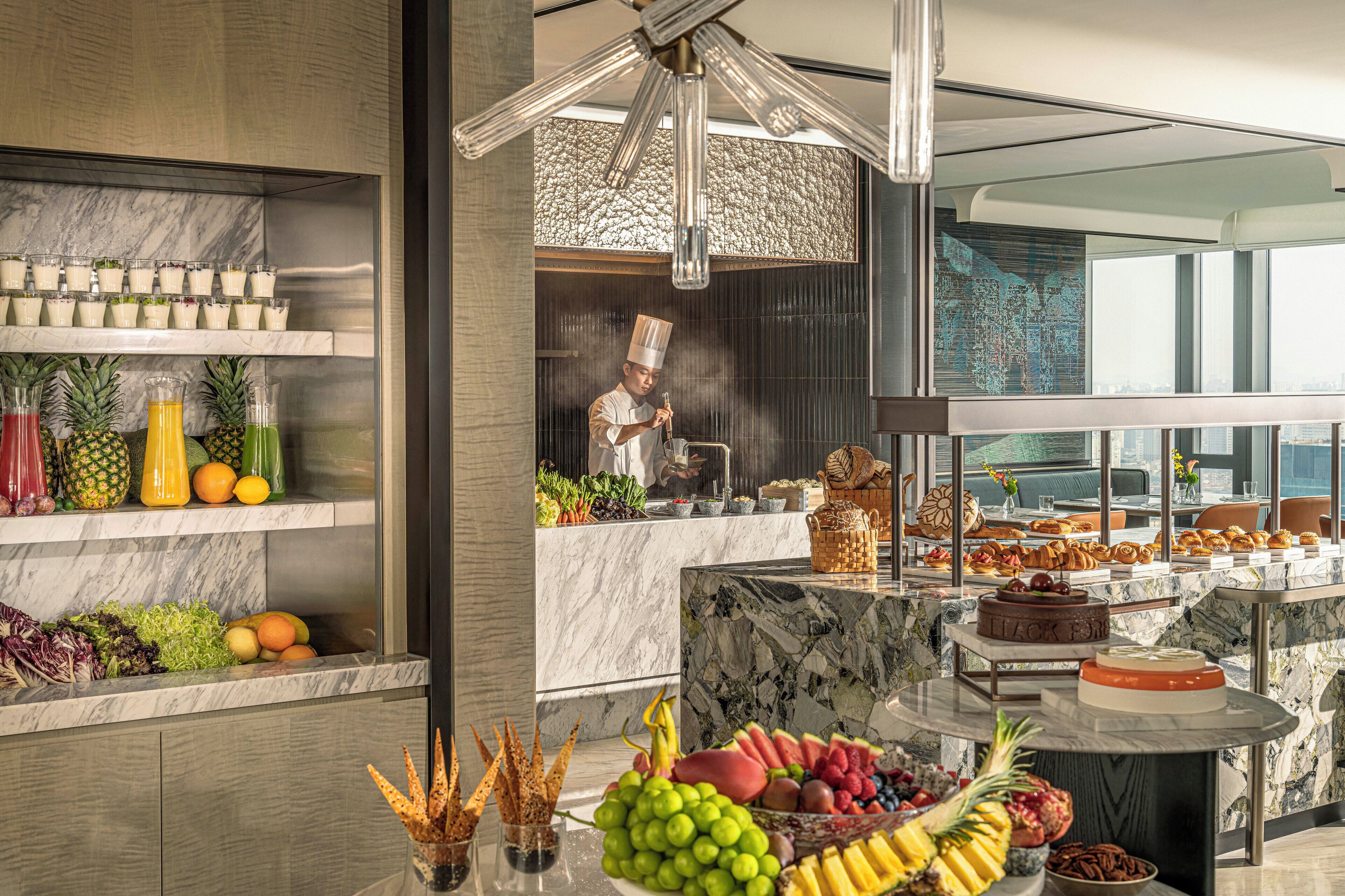 A modern kitchen with marble countertops, a chef preparing food, and a variety of fruits and pastries on display.