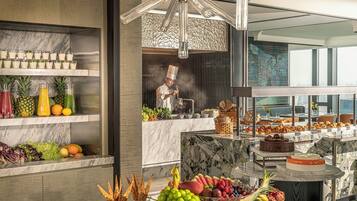 A modern kitchen with marble countertops, a chef preparing food, and a variety of fruits and pastries on display.