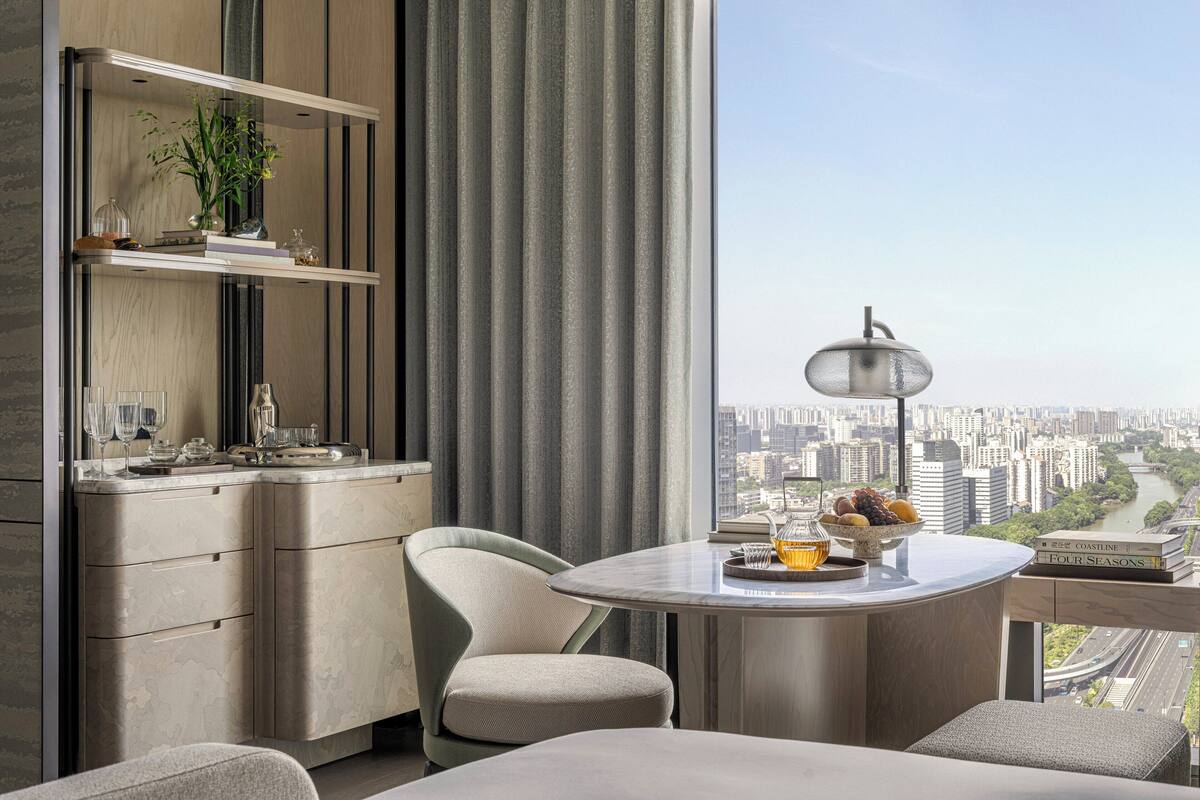 A modern hotel room with a city view, a round dining table, a comfortable chair, a minibar, and a large window with curtains.