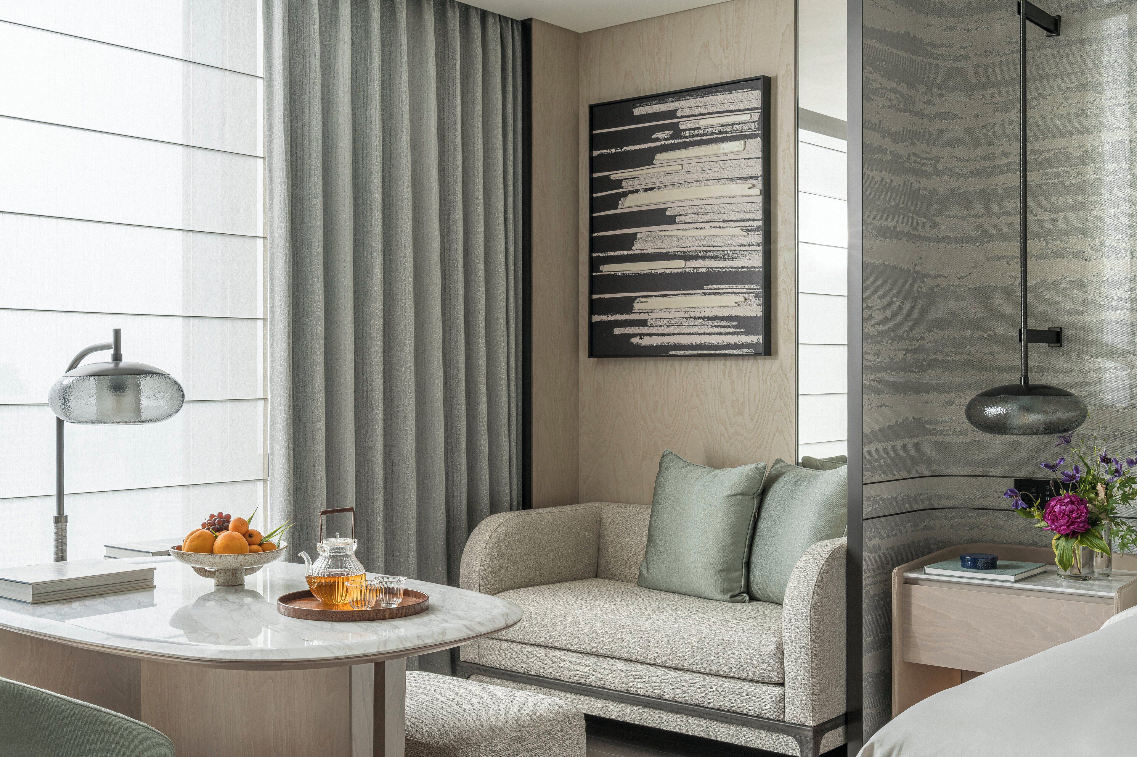 A modern hotel room with a sofa, a dining table with fruit and a teapot, a painting on the wall, and a shower area.
