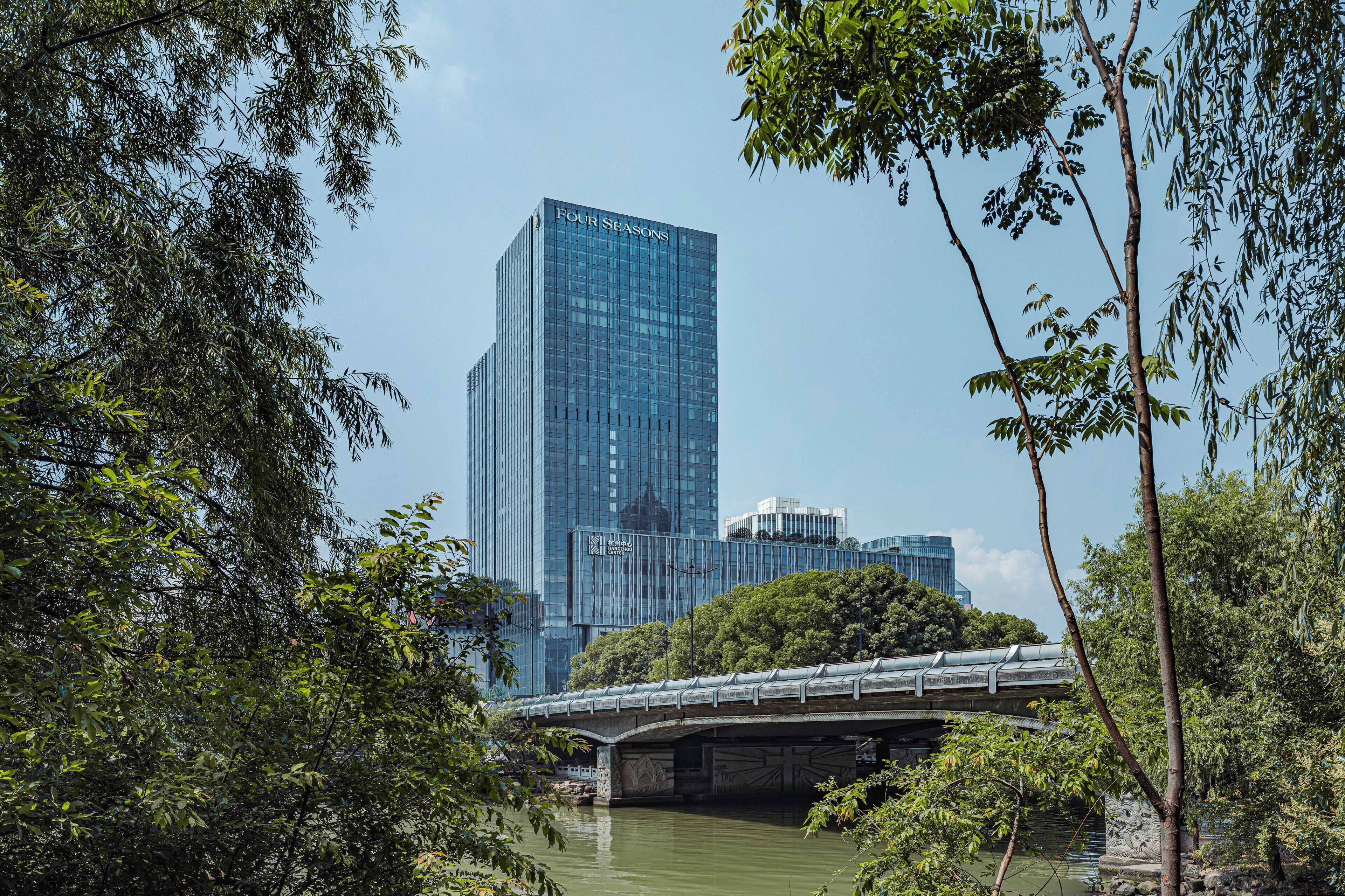 A modern building with the name "FOUR SEASONS" on its facade, surrounded by lush greenery and a bridge over a river.