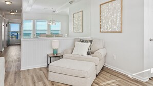 Ein Wohnzimmer mit einem beige gefärbten Sofa, einem passenden Hocker, einem kleinen Beistelltisch mit Lampe und einem großen Spiegel, der den Raum reflektiert.
