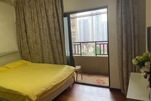 A bedroom with a bed, a small table, a window with a view of buildings, and a balcony.