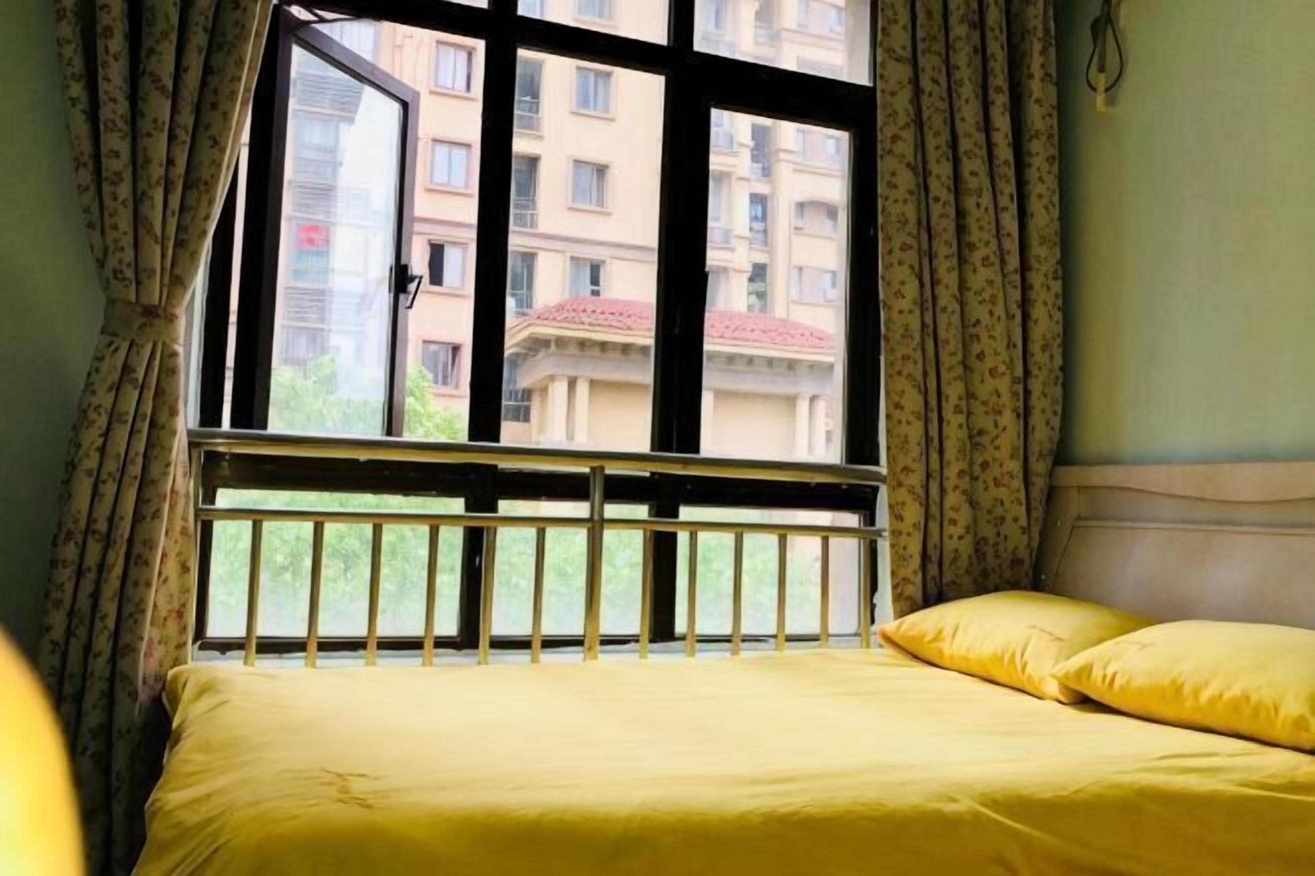 A bed with yellow bedding, a window with a view of buildings, and curtains.
