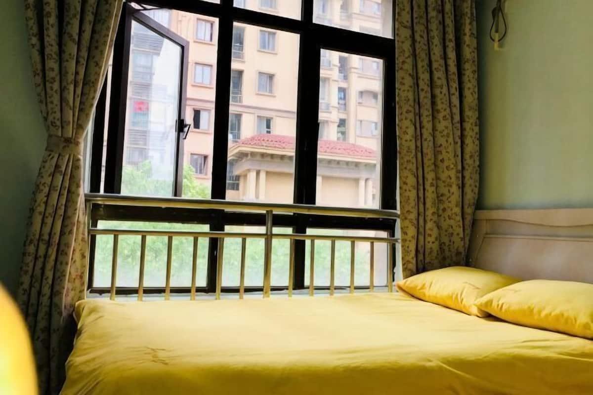 A bed with yellow bedding, a window with a view of buildings, and curtains.
