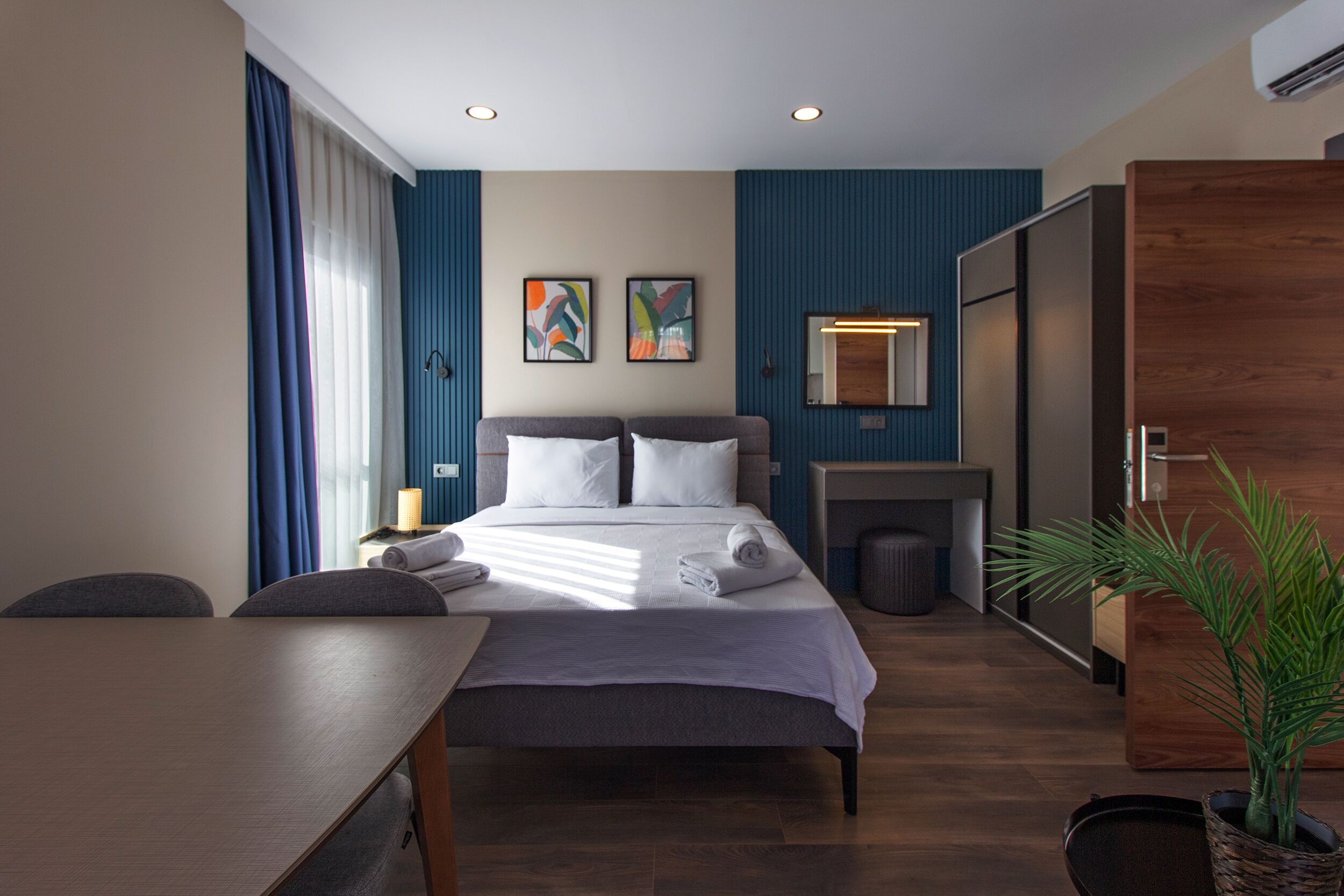 A modern hotel room with a bed, a desk, a chair, a potted plant, and a mirror.