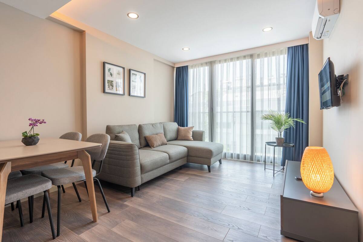 A modern living room with a sofa, dining table, and chairs. There is a TV mounted on the wall and a plant on a side table.