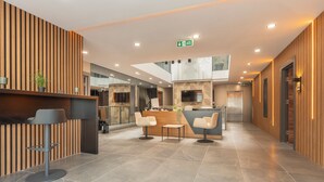 A modern reception area with a wooden reception desk, a seating area with three chairs, and a central island with a plant. - Doqu Suites Hotel (Konyaalti)