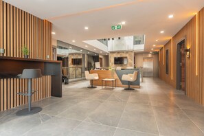 A modern reception area with a wooden reception desk, a seating area with three chairs, and a central island with a plant. - Doqu Suites Hotel (Konyaalti)