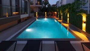 Zwembad met ligbedden en sfeerverlichting, omringd door een haag en moderne gebouwen.