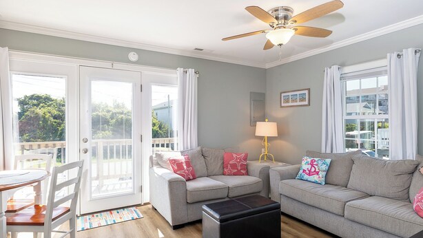 A living room with a grey sofa set, a black ottoman, a wooden dining table with chairs, and a ceiling fan. - Pelican's Nest - Soundside, 5 min to beach, 2BR 1BA, Atlantic Beach (Atlantic Beach)