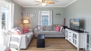 A living room with two grey sofas, a black ottoman, a ceiling fan, a TV, and a white cabinet. - Pelican's Nest - Soundside, 5 min to beach, 2BR 1BA, Atlantic Beach (Atlantic Beach)