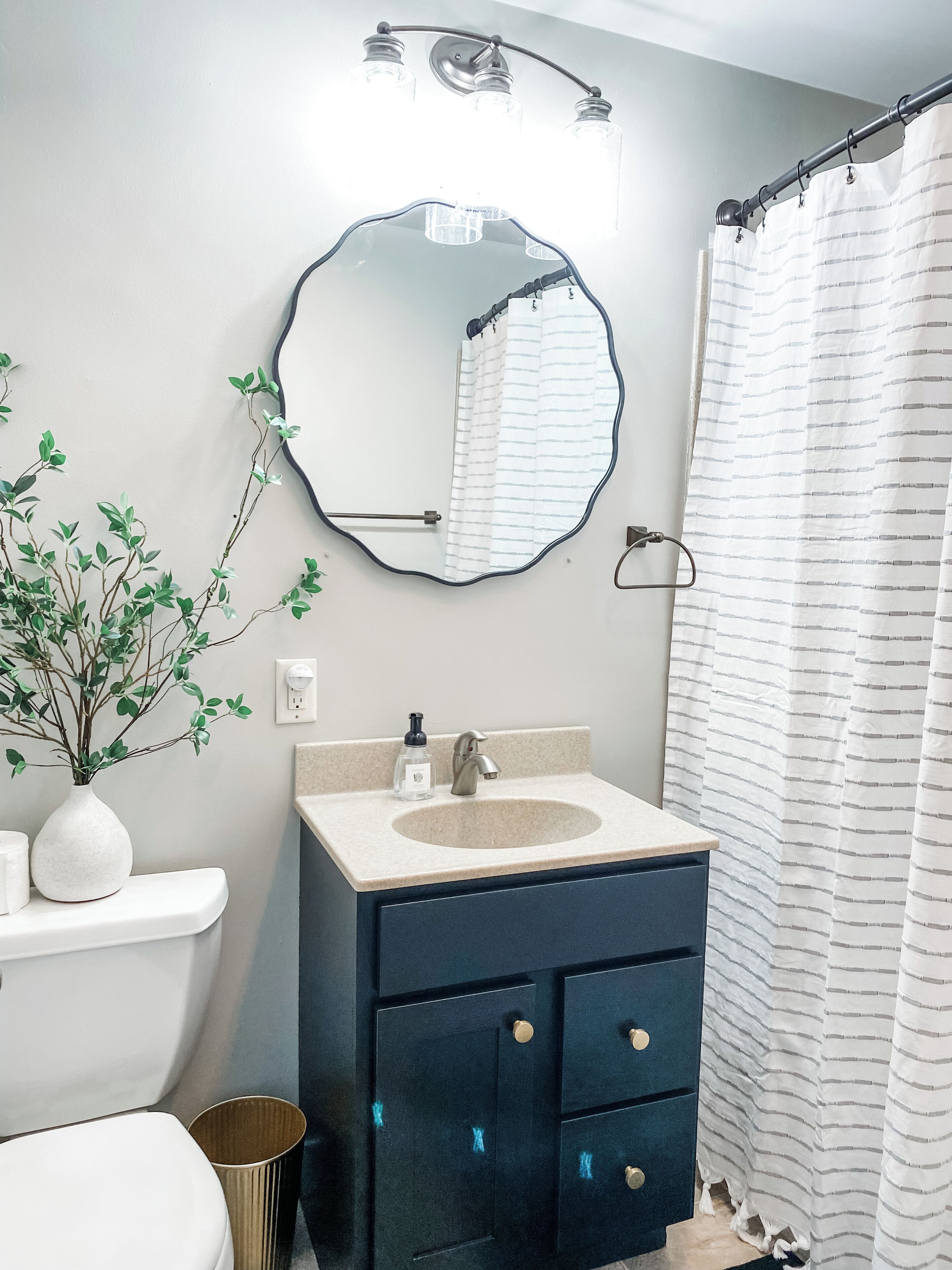 A bathroom with a white sink, dark blue vanity, a round mirror, a shower curtain, and a plant on the counter.