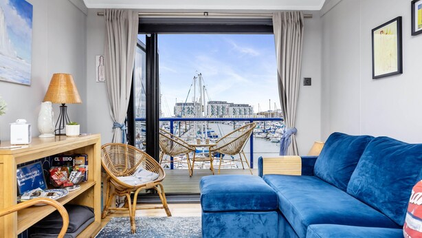 A modern living room with a blue sofa, wicker chairs, a wooden console table, and a view of a marina with boats and buildings.