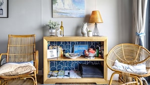 A room with two wicker chairs, a console table with books and a bottle, and a painting of a coastal scene.