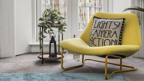 Un sillón amarillo con un cojín que dice '¡LUCES! ¡CÁMARA! ¡ACCIÓN!' junto a una mesita con una planta y un farol.
