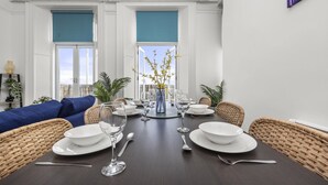 Un comedor con mesa de madera oscura dispuesta para cuatro personas, rodeado de sillas de mimbre, y vistas al cielo a través de una ventana con persianas azules.
