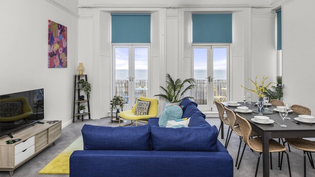 Una sala de estar moderna con un sofá azul, una mesa de comedor dispuesta para seis personas y un televisor sobre un mueble de madera.