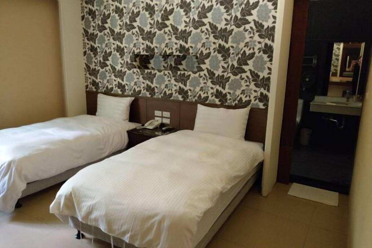 Habitación de hotel con dos camas, una mesita de noche, un espejo y un baño visible a través de una puerta abierta.