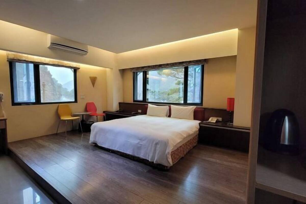 Habitación de hotel con cama, dos mesitas de noche, una silla, un escritorio y un ventanal grande.