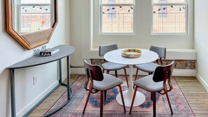 Una habitación con una mesa redonda, cuatro sillas, una pequeña consola y un espejo grande. Hay dos ventanas que dan a vistas de edificios.