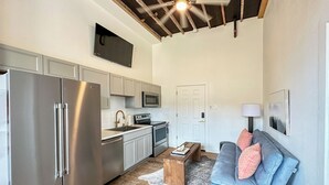 Una cocina moderna con electrodomésticos de acero inoxidable, microondas y horno empotrado. Hay una mesa de centro de madera con libros y una alfombra blanca. Un sofá azul con cojines rosas se encuentra en la sala de estar, y se ve un ventilador de techo.