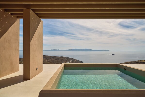 A modern infinity pool with clear water, surrounded by beige walls, overlooking a calm sea and distant land.