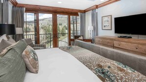 A bedroom with a large bed, a dresser, a TV, and a balcony with a view of trees and mountains.