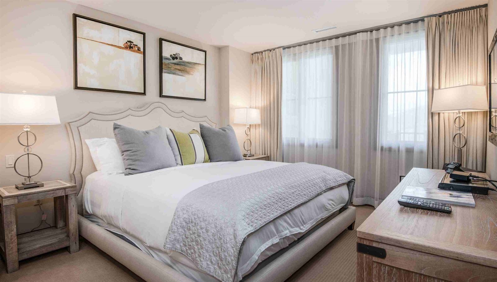 A hotel room with a large bed, bedside tables with lamps, a desk with a laptop and remote controls, and two framed pictures on the wall.