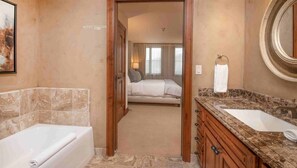 A hotel bathroom with a bathtub, a sink, and a mirror. There is a door leading to a bedroom with a bed and curtains. - Luxury 2 Bedroom Residence at The Vail Ritz-carlton (Vail)