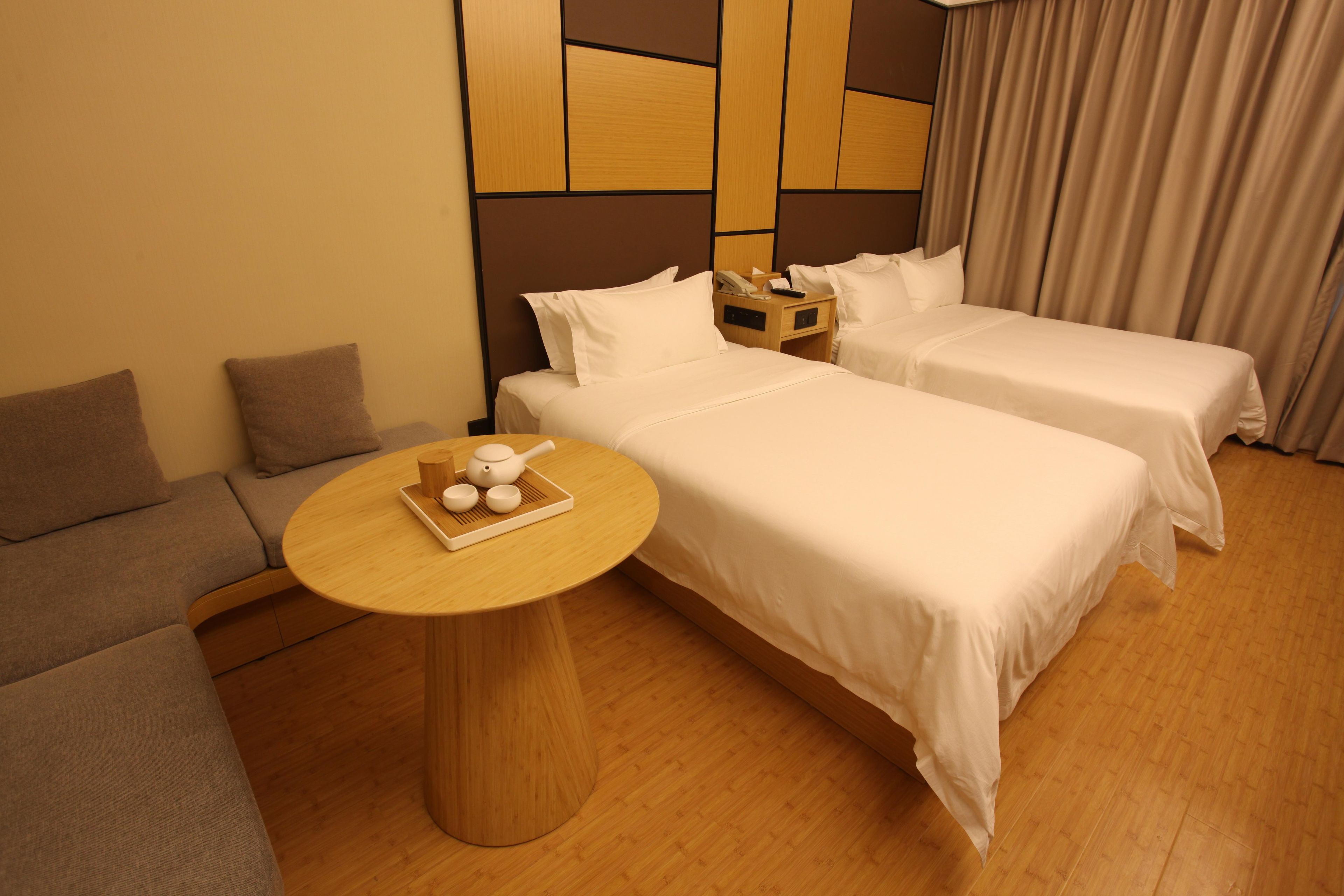 Una habitación de hotel con dos camas, un sofá, una mesa redonda de madera y una bandeja con teteras, además de una pared con paneles geométricos.