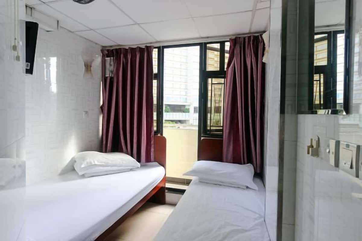 A hotel room with two beds, a window with curtains, a tiled wall, and a view of the outside.