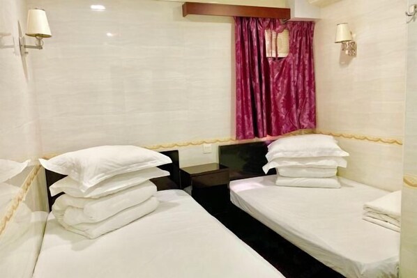 A hotel room with two single beds, a small table, a nightstand, a window with curtains, and wall-mounted lamps. - Wing Lee Guest House (Hong Kong)