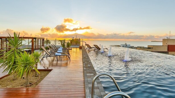 Ein Infinity-Pool auf dem Dach mit Liegestühlen und einer Holzterrasse, mit Blick auf einen Sonnenuntergang über dem Meer.