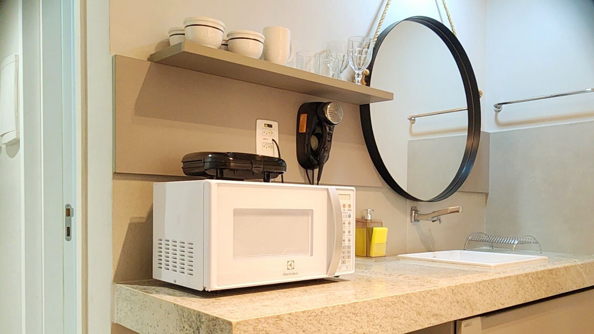 A microwave oven on a kitchen counter with a shelf above it holding cups and a hairdryer.