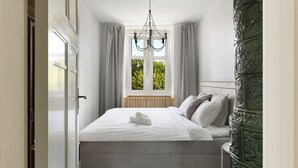 Een opgemaakt bed met een opgerolde handdoek, een raam met gordijnen en een kroonluchter.