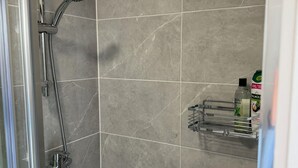 Zona de ducha con puerta de vidrio, baldosas de mármol gris y un estante con artículos de aseo.